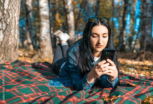Young beautiful woman looks into the phone, writes a message while lying on a blanket in an autumn park, chatting on the Internet. The student is resting in the park after school. Autumn time