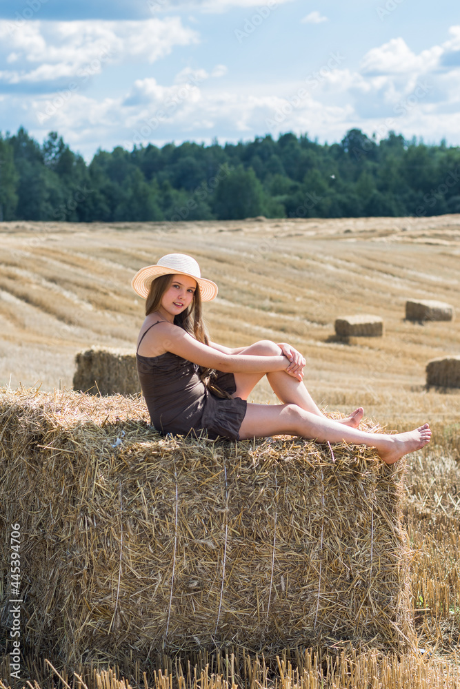A beautiful teenager girl in a hat sits in a field on a haystack on a sunny day. A beautiful teenager girl in a hat sits in a field on a haystack on a sunny day.