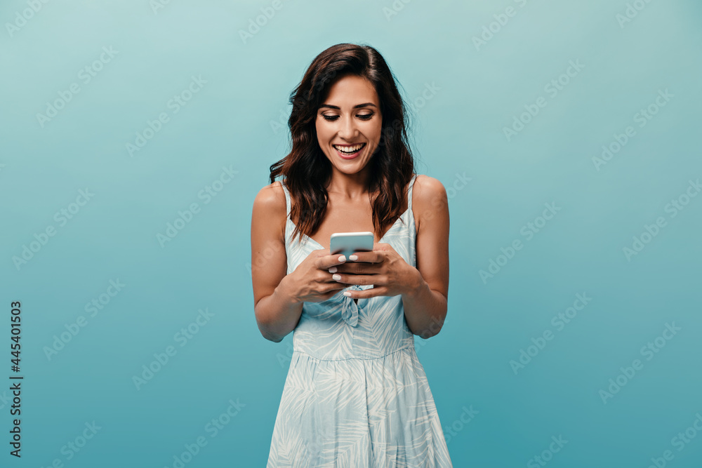 Mischievous girl in white dress with smile chatting in smartphone on blue background. Cheerful lovely woman in good mood holds phone Mischievous girl in white dress with smile chatting in smartphone on blue background. Cheerful lovely woman in good mood holds phone