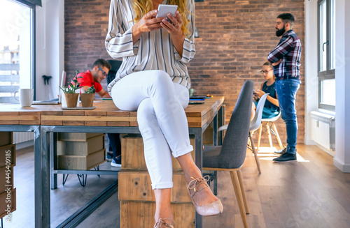 Unrecognizable businesswoman sitting on table looking mobile in the office with colleagues working in the background photo