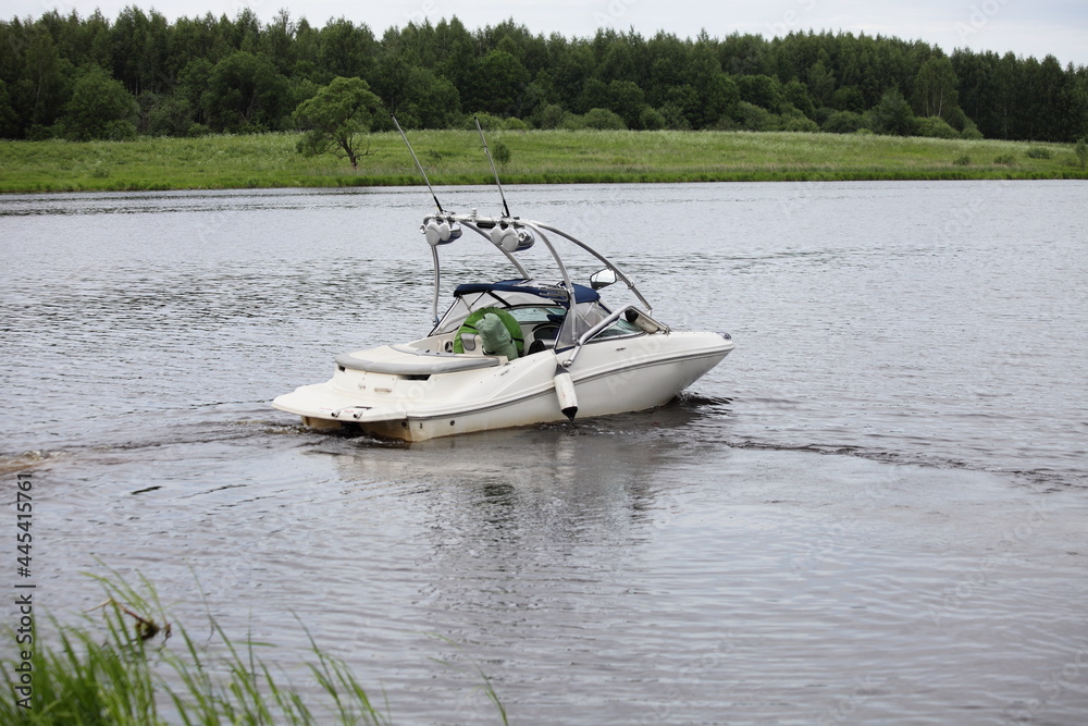 Obraz premium White powerboat with towing targa slowly float on river calm water on green grassy shore with forest background at summer day, front view, active outdoor recreation watersports in Europe