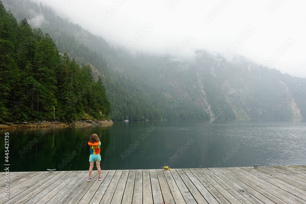 A little girl with her life jacket on standing on a dock admiring the view of the misty princess louisa inlet and surrounding forest, in British Columbia, Canada. A little girl with her life jacket on standing on a dock admiring the view of the misty princess louisa inlet and surrounding forest, in British Columbia, Canada.