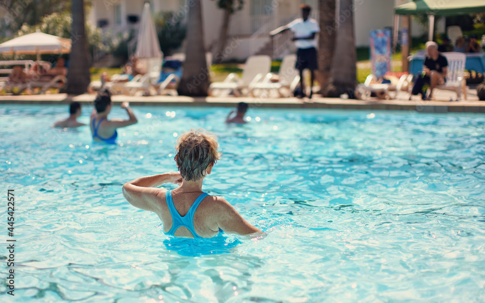 Elderly senior woman with grey hair, wearing blue swimsuit doing water aerobics in hotel pool, view from behind