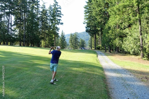 An older male swinging his club from the rough on an approach shot on a par 4 surrounded by forest, on a beautiful sunny day in Port Alberni, British Columbia, Canada. photo