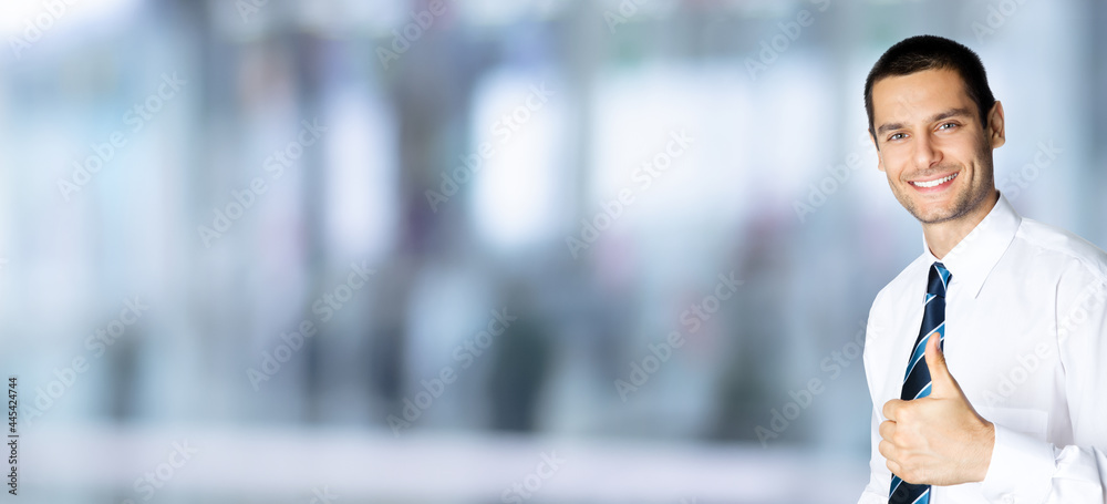 Portrait of smiling businessman in white shirt and tie, showing thumb up like hand sign gesture, over blurred modern office interior background. Happy confident man gesturing. Success in business.