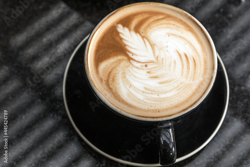Simple dramatic overhead shot of a decorated cappuccino. Foam with a beautiful pattern by a barista. In a black mug, on a black saucer, on a black granite surface. Reflection of a pergola on surface