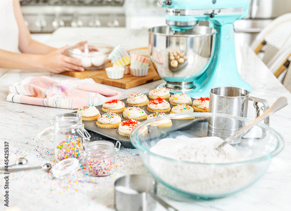 Pastel kitchen scene with cupcakes. Teal mixer sits with a cupcake baking tray full of cupcakes decorated with red and multicolor sprinkles. Hand placing cupcake on left side. . Pastel kitchen scene with cupcakes. Teal mixer sits with a cupcake baking tray full of cupcakes decorated with red and multicolor sprinkles. Hand placing cupcake on left side. .