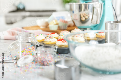 Pastel kitchen scene with cupcakes.  Teal mixer sits with a cupcake baking tray full of cupcakes decorated with red and multicolor sprinkles.  Foreground has sugar and flour and jars with sprinkles.