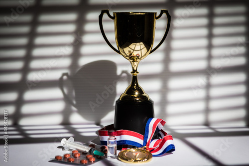 Gold medal and champion's cup, syringe with doping substance, pill tablet and vial with prohibited substance with lights and shadows of a curtain entering through the window. Sport and doping concept