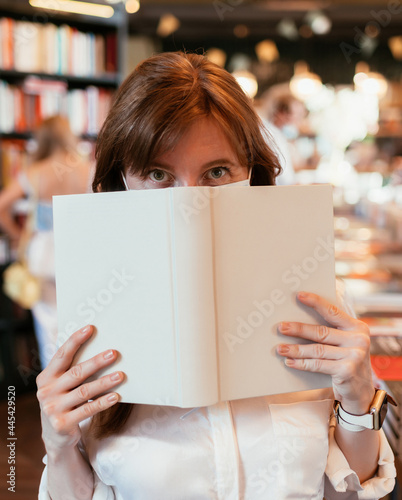 Girl in a medical mask in the library covers half of her face with a book