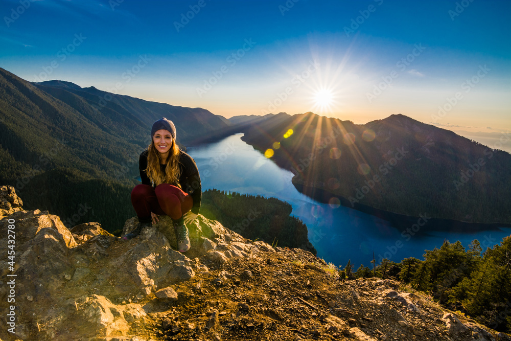 Adventurous athletic woman hiker sitting on a mountain top smiling at the camera with a beautiful sunset over mountains and a lake.