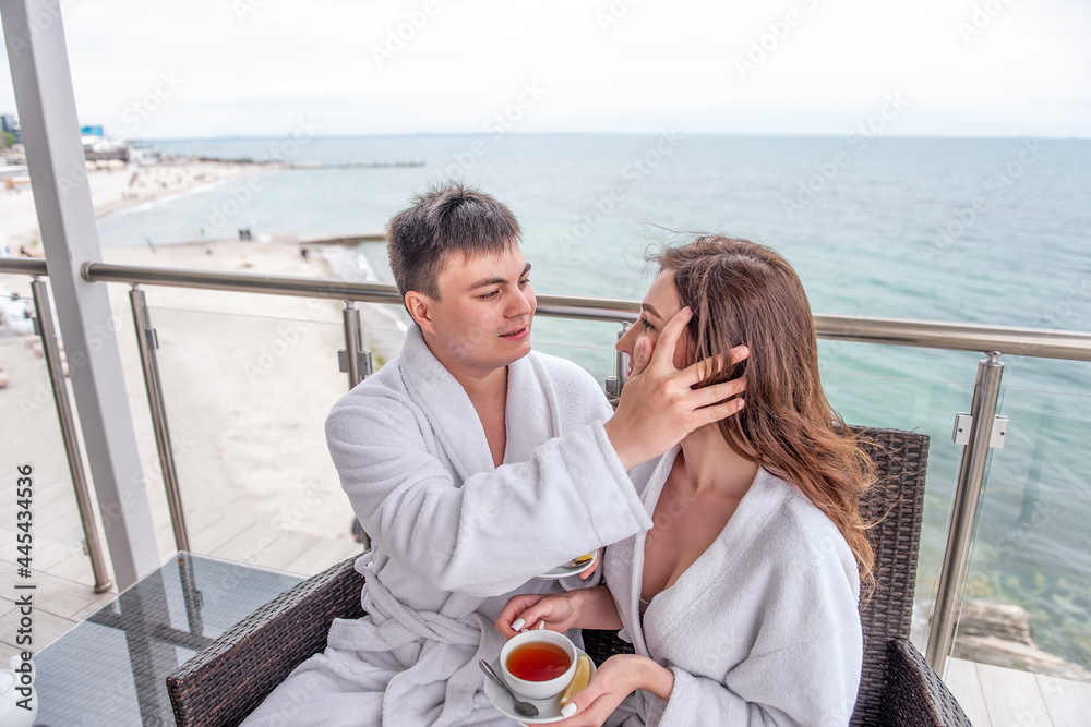 Couple in white bathrobes are sitting on an open terrace by the sea, drinking tea with lemon from cups. Lovers travel on vacation. A young man straightens a woman's hair. Newlyweds on honeymoon