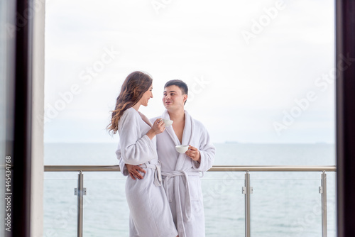 Young loving couple in white terry robes drink tea from cups, the background of blue sea. Newlyweds spend honeymoon traveling. Lovers in love are happy laughing hugging, looking at each other. Resting