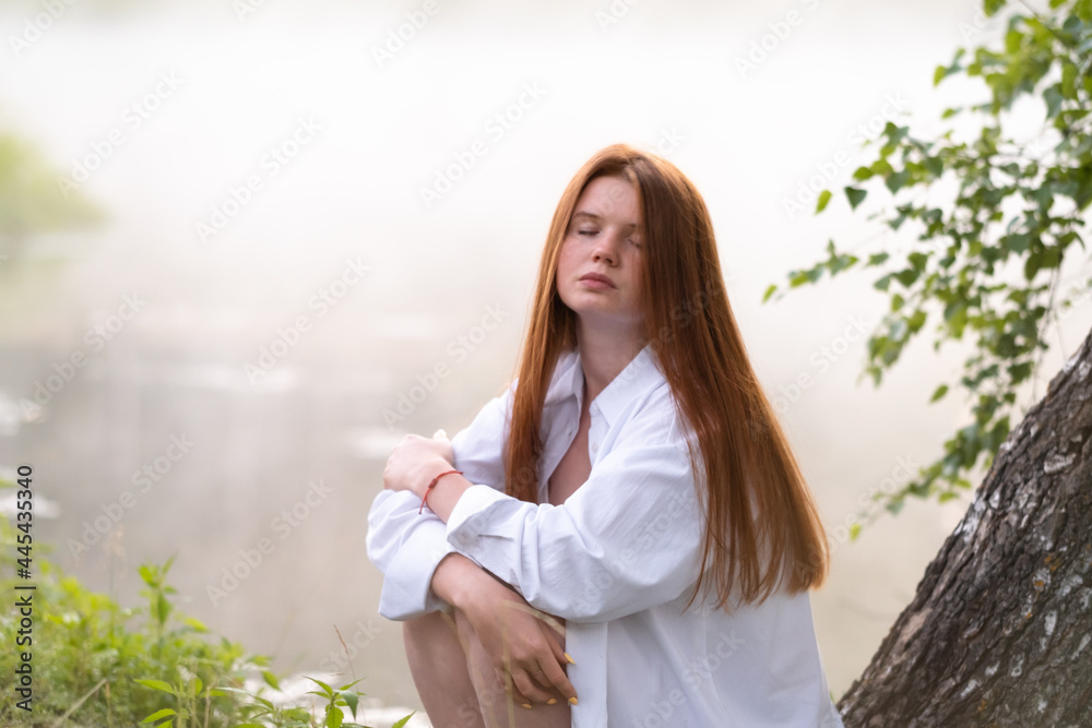 Fototapeta premium A beautiful red-haired girl in a white shirt sits by the lake on a tree in the morning fog. Meeting the dawn in nature.