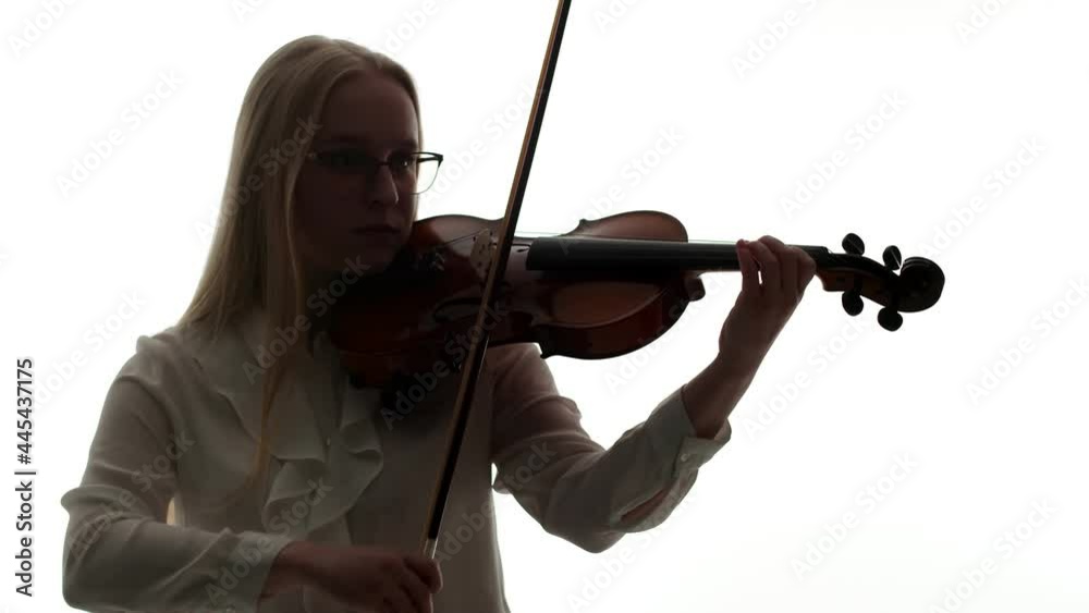 Blonde woman in glasses and white blouse plays with bow and fingers on strings of the violin in dark light of the studio. Artist is a woman engaged a string musical instrument, white background. Blonde woman in glasses and white blouse plays with bow and fingers on strings of the violin in dark light of the studio. Artist is a woman engaged a string musical instrument, white background.