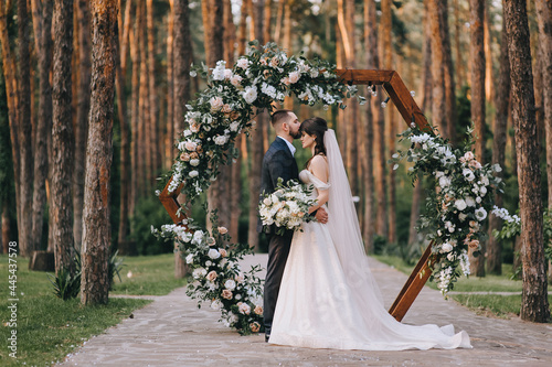 Stylish groom in a suit and a cute brunette bride in a white dress in the forest near a wedding wooden arch decorated with flowers. Wedding portrait of the newlyweds. photo