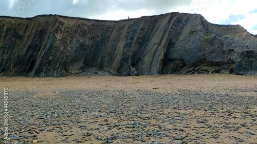 Tall slate rocks aligned vertically forming a cliff a few metres high in Widemouth beach in Conrwall, England. Some people seen walking along the top of it. The beach sand is mostly cover by pebbles. photo