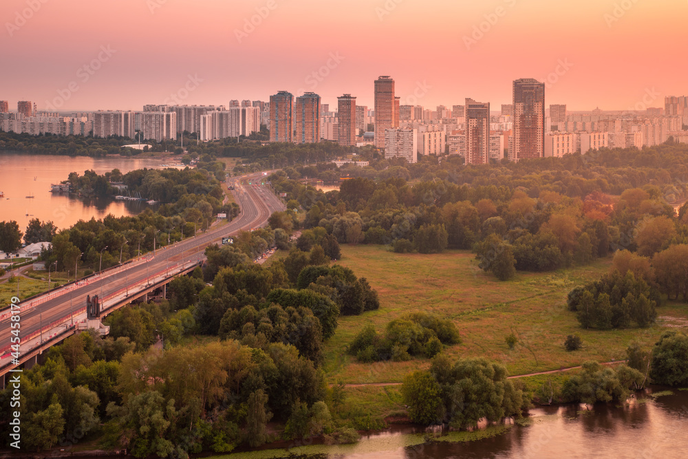 Aerial view of a summertime sunset cityscape featuring an s-curved highway leading to a residential area of a large city against a clear pink sky