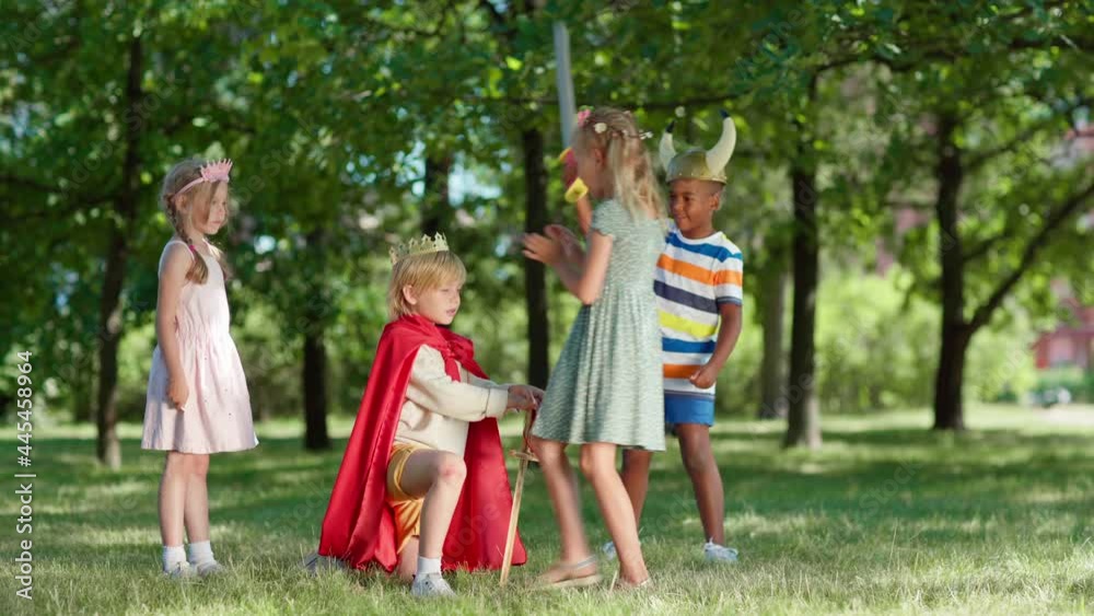 Diverse kids in costumes having fun role playing in park. Little boy in red cape being coronated kneeing on grass with sword in his hand. Boy standing up, crown falling. Children applauding to king Diverse kids in costumes having fun role playing in park. Little boy in red cape being coronated kneeing on grass with sword in his hand. Boy standing up, crown falling. Children applauding to king