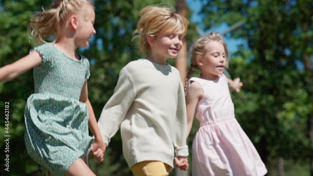 Tracking side view shot of group of three joyful blonde kids, boy and two girls, running holding hands. Friendly children hopping in park in slow motion feeling happy and carefree Tracking side view shot of group of three joyful blonde kids, boy and two girls, running holding hands. Friendly children hopping in park in slow motion feeling happy and carefree