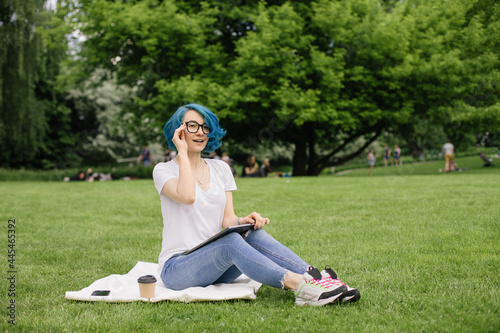 Young women with blue hair sitting on green grass with laptop in the hands.