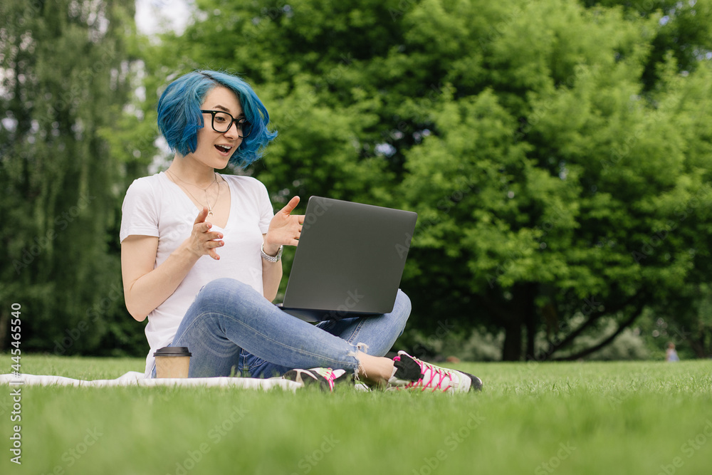 Young women with blue hair sitting on green grass with laptop in the hands.