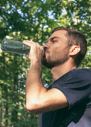 Adult man drinking water from reusable plastic bottle after sport in nature. Recovery, refreshing, healthy lifestyle.