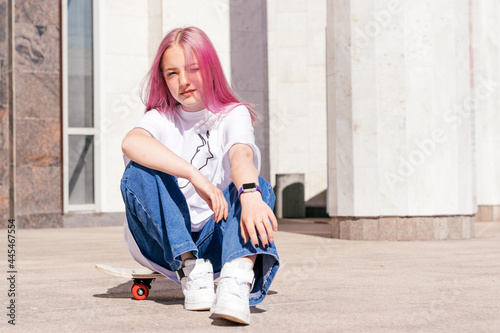 A pink-haired hipster teenage girl in a white T-shirt and jeans is sitting on a skateboard on a city street.Generation Z style,active lifestyle,summer concept.Copy space for text.
