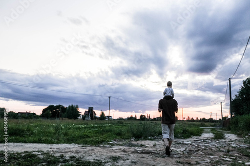 Father with his son sitting on his neck goes into the sunset, to the field, into the distance, evening light, frontal photography, green background, sunset, general plan