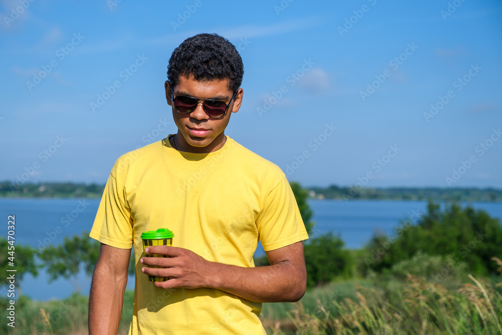 Smiling African American man in sunglasses enjoying the taste of coffee outdoors in summer by the river, looking and standing in front of the camera. Copy space