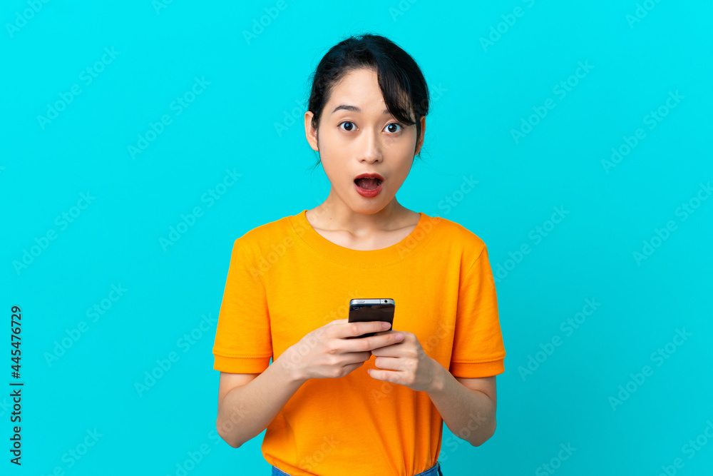 Young Vietnamese woman isolated on blue background looking at the camera while using the mobile with surprised expression Young Vietnamese woman isolated on blue background looking at the camera while using the mobile with surprised expression