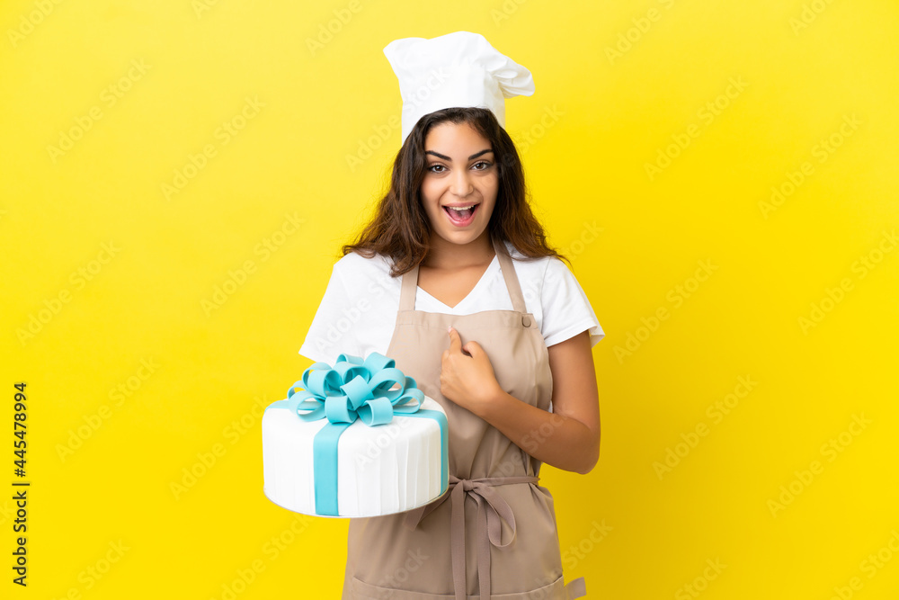 Young caucasian pastry chef woman with a big cake isolated on yellow background with surprise facial expression Young caucasian pastry chef woman with a big cake isolated on yellow background with surprise facial expression