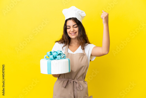 Young caucasian pastry chef woman with a big cake isolated on yellow background celebrating a victory