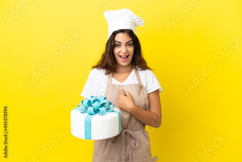 Young caucasian pastry chef woman with a big cake isolated on yellow background with surprise facial expression