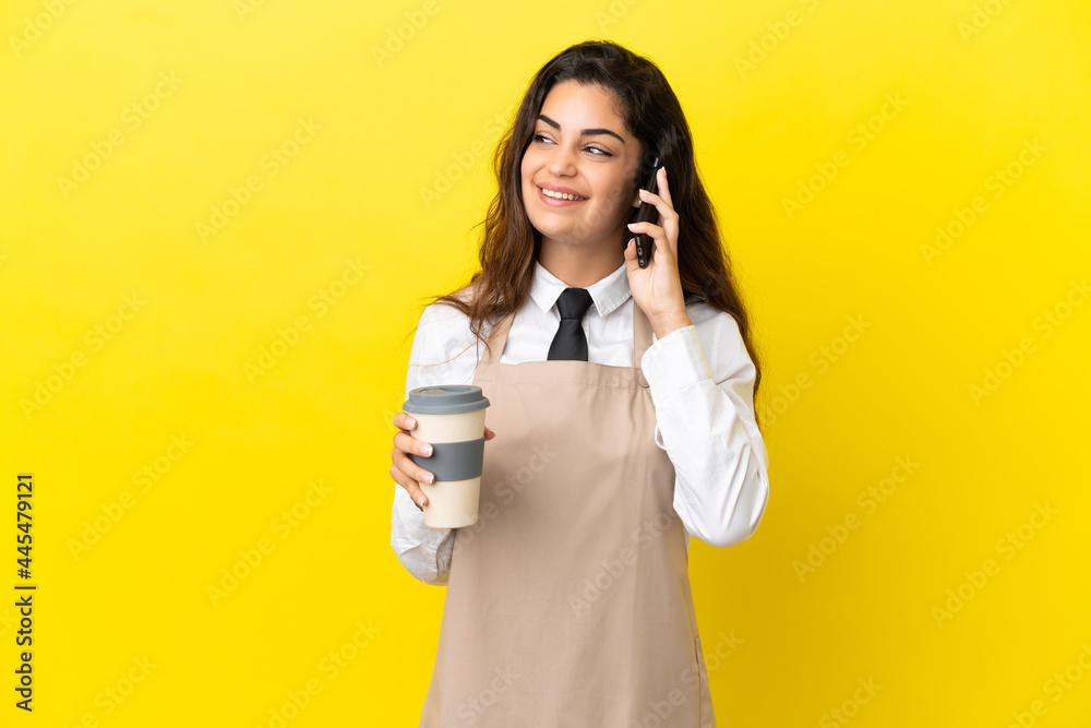 Young caucasian restaurant waiter isolated on yellow background holding coffee to take away and a mobile Young caucasian restaurant waiter isolated on yellow background holding coffee to take away and a mobile