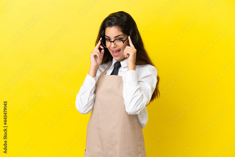 Young caucasian restaurant waiter isolated on yellow background with glasses and surprised Young caucasian restaurant waiter isolated on yellow background with glasses and surprised