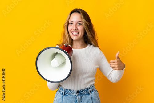 Young blonde woman isolated on yellow background holding a megaphone with thumb up