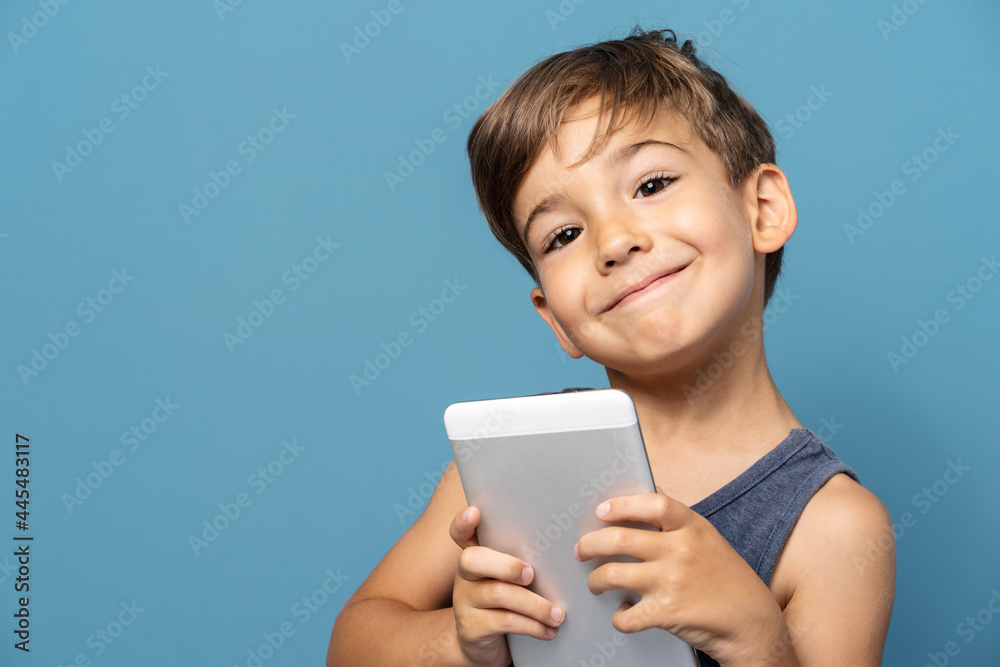 Front view of small caucasian boy four years old standing in front of blue background studio shot standing and using digital tablet looking to the camera happy smiling Front view of small caucasian boy four years old standing in front of blue background studio shot standing and using digital tablet looking to the camera happy smiling