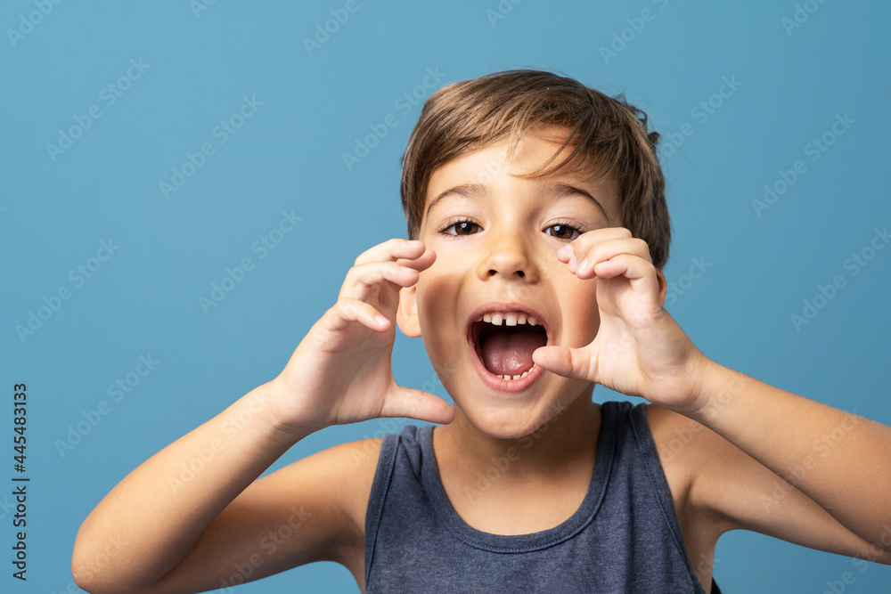 Front view of small caucasian boy four years old standing in front of blue background studio shot standing and holding hands in front of his opened mouth like he is shouting Front view of small caucasian boy four years old standing in front of blue background studio shot standing and holding hands in front of his opened mouth like he is shouting