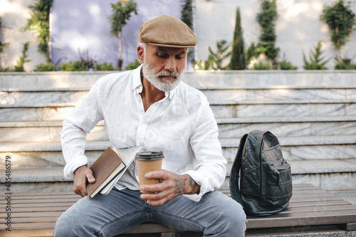 Adult man in trendy outfit posing with laptop and notebooks outside. Serious modern guy with white beard is sitting on bench next to black briefcase..