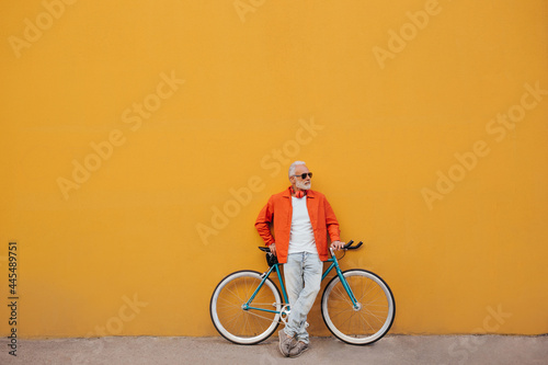 Stylish man in sunglasses leans on bicycle on orange wall background. Gray-haired fashionable guy in jeans, white T-shirt and in bright jacket poses..