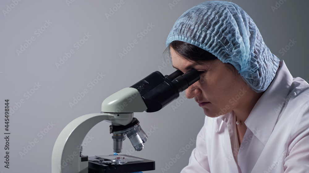 Skilled lady lab assistant in cap and white coat looks at material sample through modern microscope at workplace in clinic laboratory copy space Skilled lady lab assistant in cap and white coat looks at material sample through modern microscope at workplace in clinic laboratory copy space