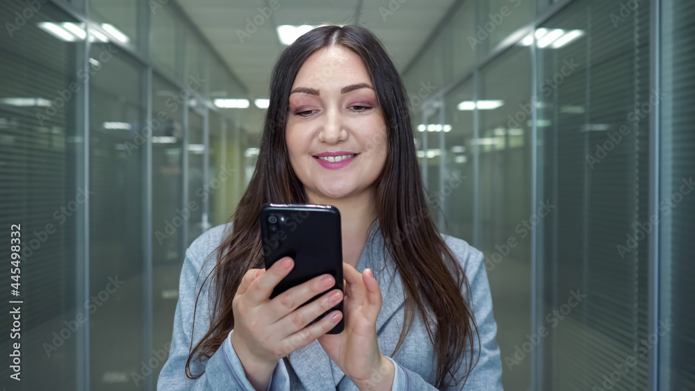 Successful lady company worker looks into smartphone display typing and smiling in office building hallway under electric light in evening closeup