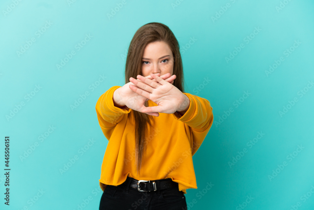 Teenager girl over isolated blue background making stop gesture with her hand to stop an act Teenager girl over isolated blue background making stop gesture with her hand to stop an act