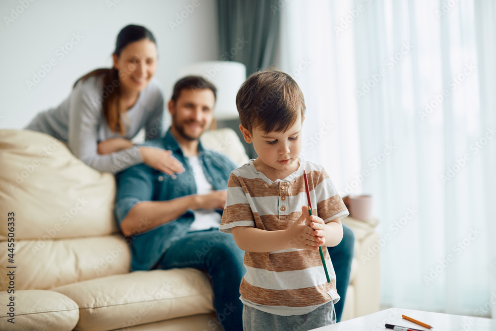 Cute boy uses crayons and enjoying in creative time at home. His parents are in the background. Cute boy uses crayons and enjoying in creative time at home. His parents are in the background.