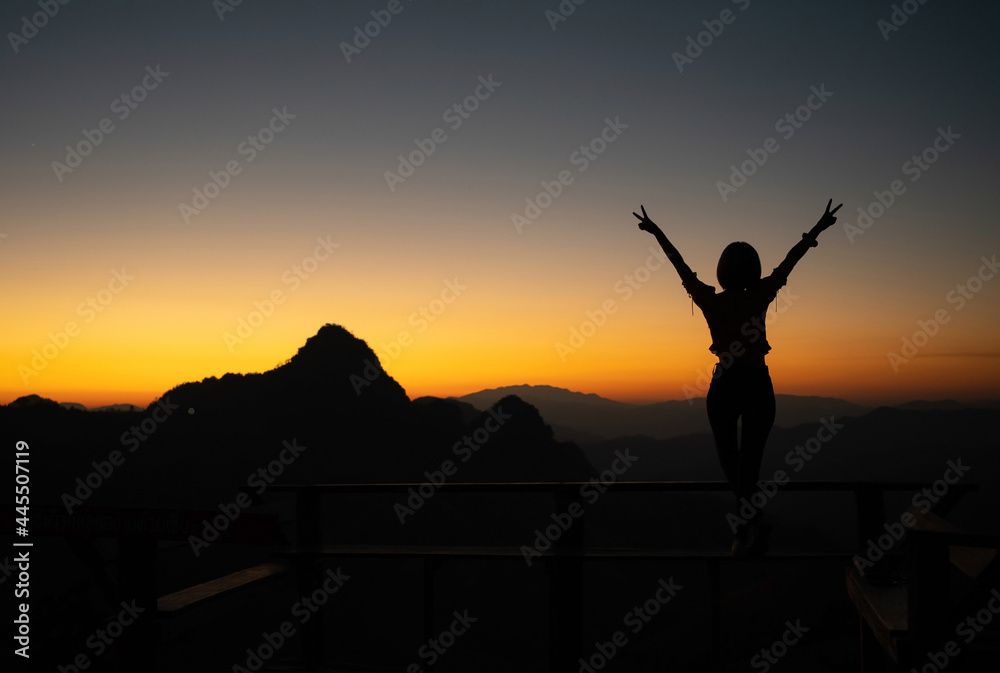 Silhouette of young Asian woman standing on a bench with raising two fingers on the sky while admires the fantastic landscape of mountain peaks with a soft golden horizon line on sunset or sunrise