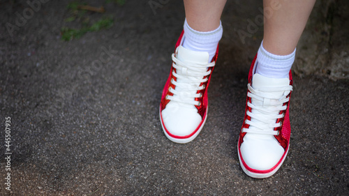 Photo of female legs in red and white sneakers on gray wet asphalt. Red and white sneakers with white laces close up