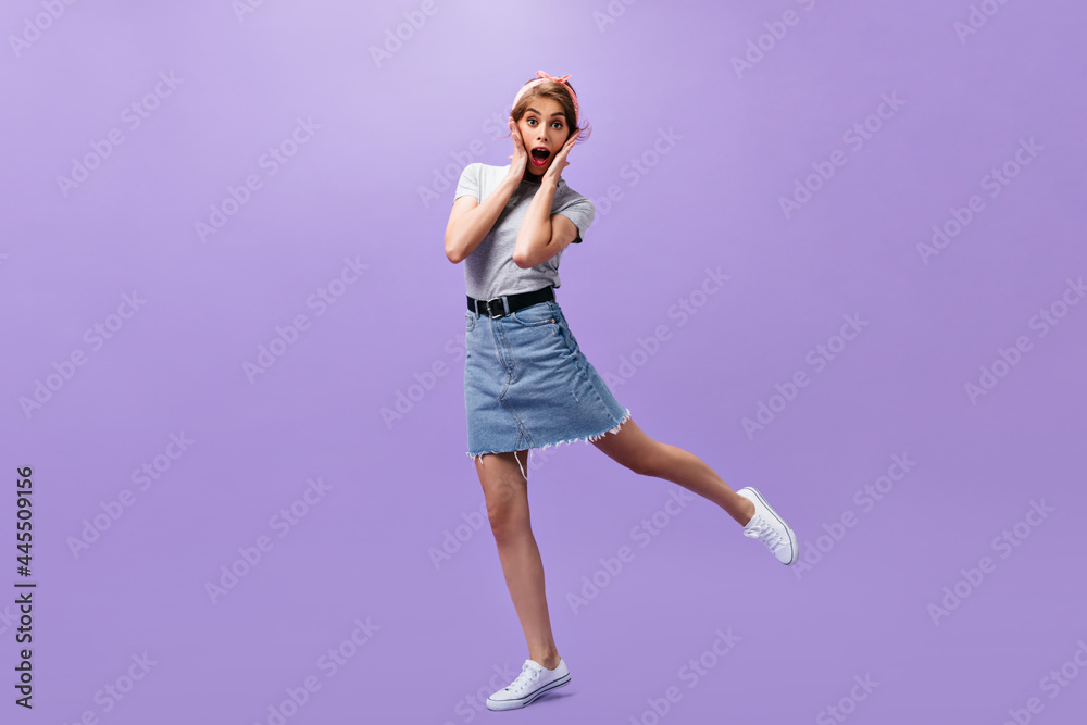 Shocked woman in stylish outfit jumps on purple background. Surprised modern lady in grey t-shirt and denim short skirt posing.. Shocked woman in stylish outfit jumps on purple background. Surprised modern lady in grey t-shirt and denim short skirt posing..