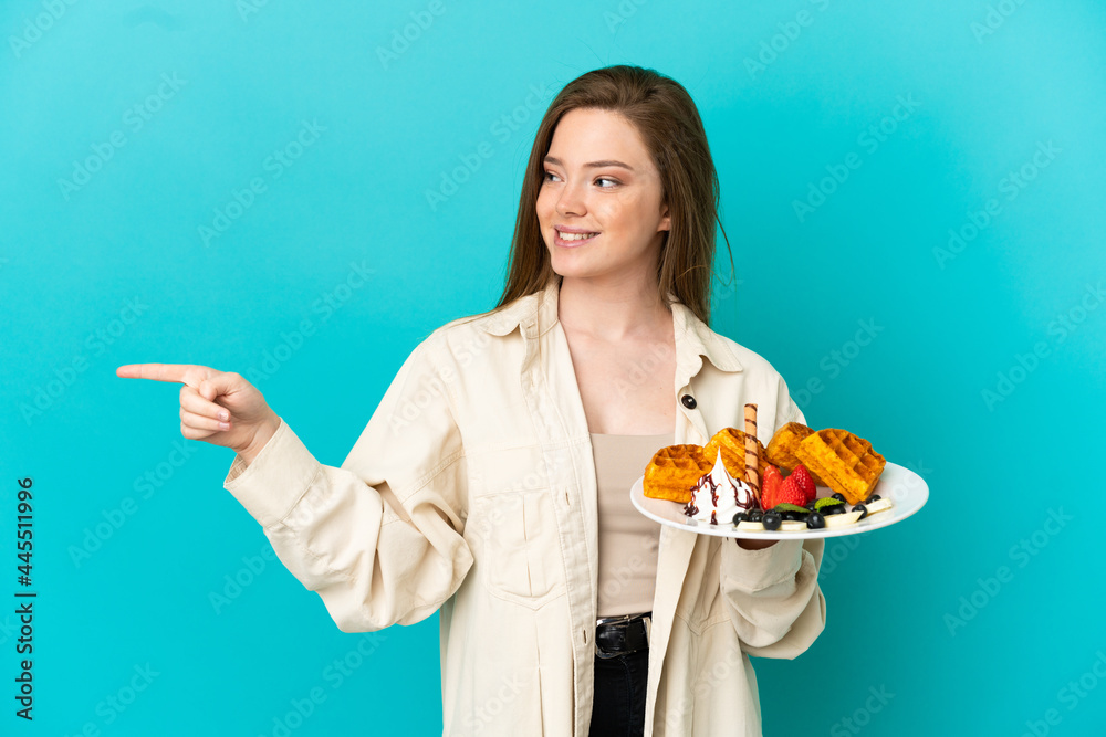 Teenager girl holding waffles over isolated blue background pointing finger to the side and presenting a product Teenager girl holding waffles over isolated blue background pointing finger to the side and presenting a product