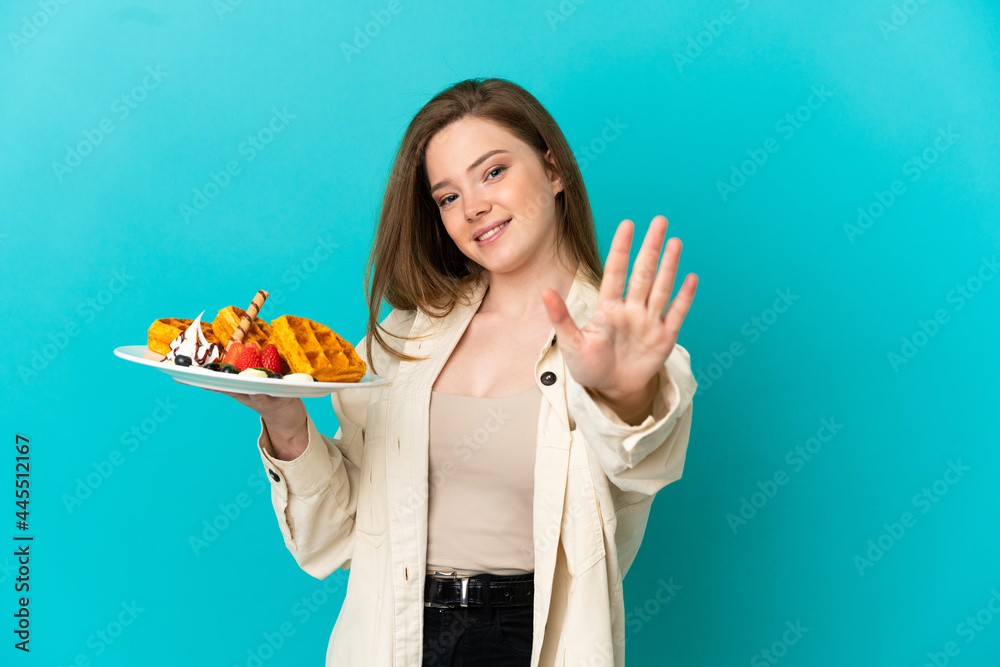 Teenager girl holding waffles over isolated blue background counting five with fingers Teenager girl holding waffles over isolated blue background counting five with fingers